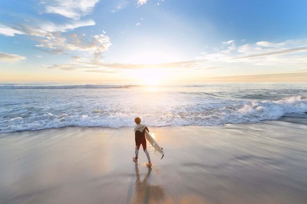 A surfer walking into gentle surf on a wide Southern California beach